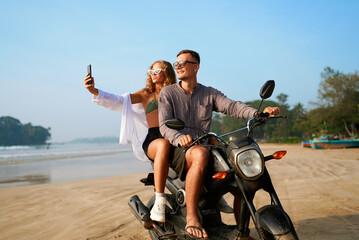 Happy couple on scooter beach ride. Smiling girl takes selfie, adventurous travel content. Young...
