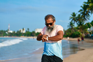 Senior runner man uses smart watch at ocean beach. Bearded male with earbuds, modern fitness tech. Healthy training, wellbeing, health check. Wireless gadget for workout tracking. Business, leisure.