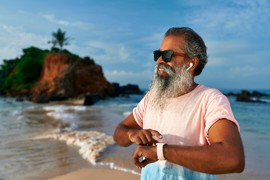 Senior bearded man runs at beach uses fitness tracker watch. Grandpa with wireless earbuds listens music. Modern gadget for health check. Old male enjoys active lifestyle. Tech helps wellbeing.