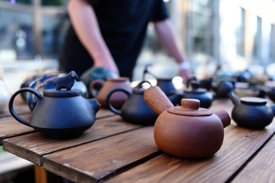 Clay teapot and handmade dishes on wooden table. Traditions of Ukrainian pottery. Man in black apron stands next to tray of clay dishes. Sale of handmade goods at art festival. Tea set for kitchen.