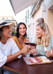 Vertical. Three happy female friends are toasting with glasses of red wine while sitting at a table outside a restaurant in a picturesque Italian town, enjoying a selection of cheese and cured meats