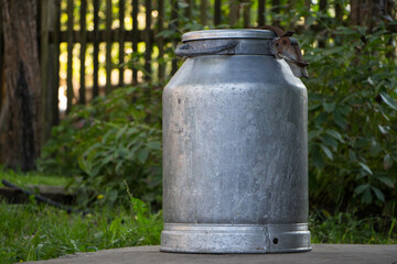 Naklejka premium Vintage aluminum milk can standing outdoors against a rustic fence and green foliage, capturing a nostalgic rural scene and a sense of simpler times