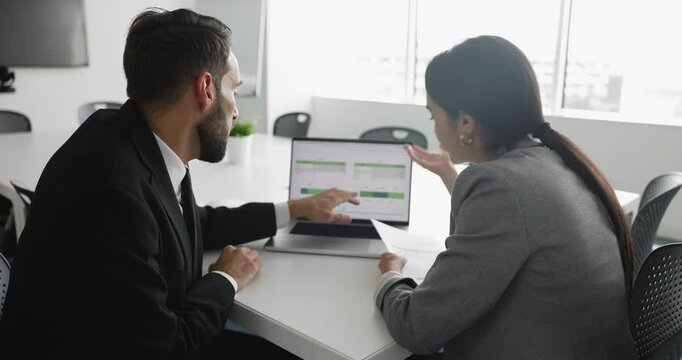 Two professionals discuss data displayed on laptop screen, reviewing performance metrics, financial data, analytical dashboards, colleague explaining or highlighting specific points shown on charts
