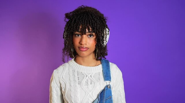 Young woman in white winter earmuffs confidently posing against vibrant purple background, showcasing contemporary fashion style and cheerful expression