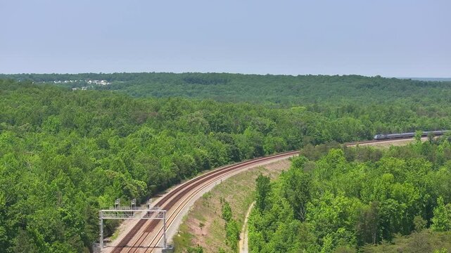 Daytime overhead view from a drone of two parallel railroad train tracks and a passenger train near trees and an industry in a rural location.