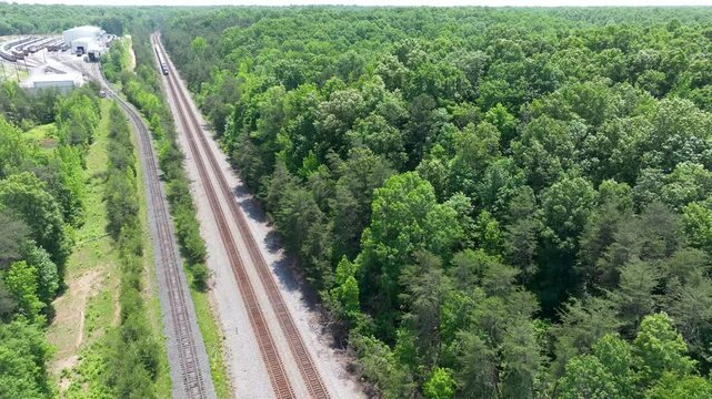 Daytime overhead view from a drone of two parallel railroad train tracks and a commuter passenger train near fields and industry in a rural location.