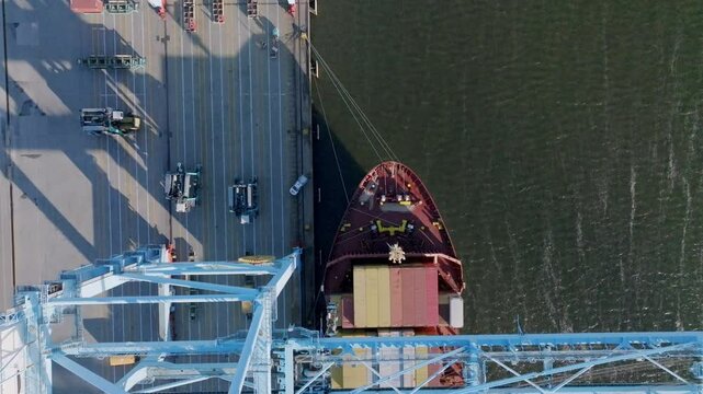 Daytime topdown view moving rearward aft from a drone of a large container freight ship being loaded and unloaded by a crane while moored at pier.
