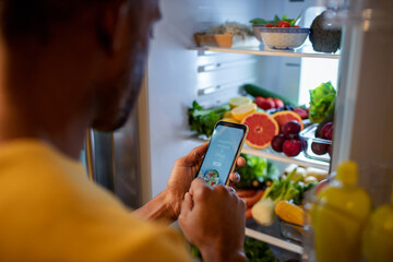 Man looking up healthy recipes on smartphone in front of open fridge with fresh vegetables