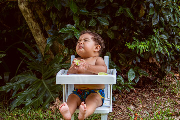9-month-old baby eating lemon in his white highchair, outdoors.
