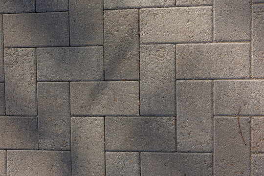 A small twig lies on interlocking rectangular pavers forming a herringbone pattern in a coarse brick walkway. Subtle shadows create a play of light and texture across the surface.