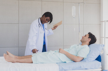 Obraz premium Woman doctor black people stand with patient caucasian man lying bed carer physical therapist two person talk help support and check treat sick person take care of your health in room hospital clinic
