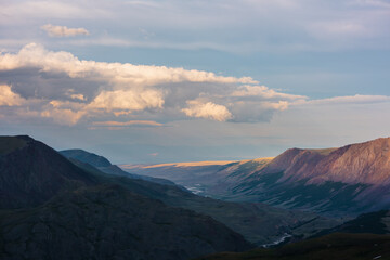 Dramatic aerial top view to golden steppe illuminated by setting sun and mountain range silhouette under clouds in gold sunset tones. Evening alpine landscape under cloudy sky in vivid sunset color.