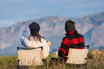A couple sits on camping chairs beside a lake, enjoying hot drinks while facing a scenic mountain backdrop. Dressed in warm clothes and beanies, they relax in the crisp outdoor morning air.