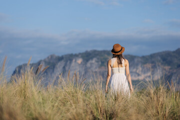 A young woman in a white dress and hat stands in a field of tall grass, gazing at the distant...