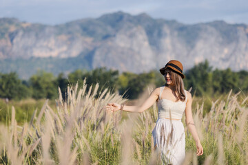 A young woman in a white dress and hat walks through a field of tall grass, gazing downward in a...