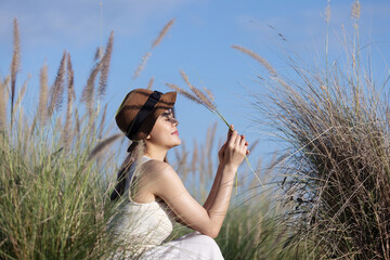 A young woman in a white dress and hat sits among tall grass, holding a stalk and gazing into the...
