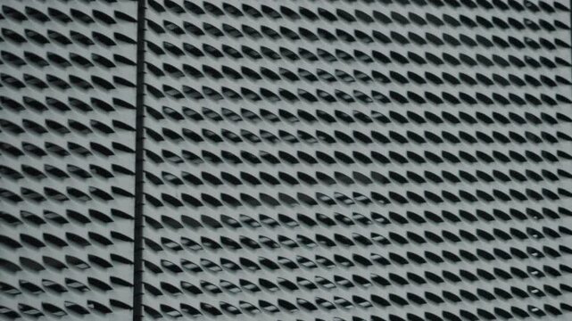 Close-up of a modern building's facade with a repetitive geometric metal screen pattern. The gray metallic texture creates an abstract design with light and shadow interplay. Ideal for architectural
