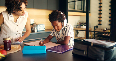 Mother, homework and child learning with headphones in kitchen for education podcast and math assessment. African family, student and books with lesson support, knowledge development and study music
