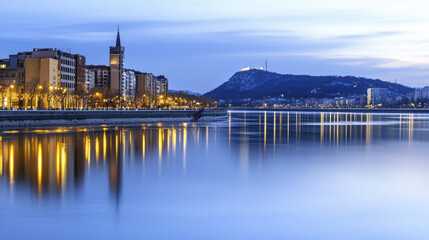 Fototapeta premium serene cityscape at dusk, featuring illuminated buildings along calm river, with distant hill and castle in background
