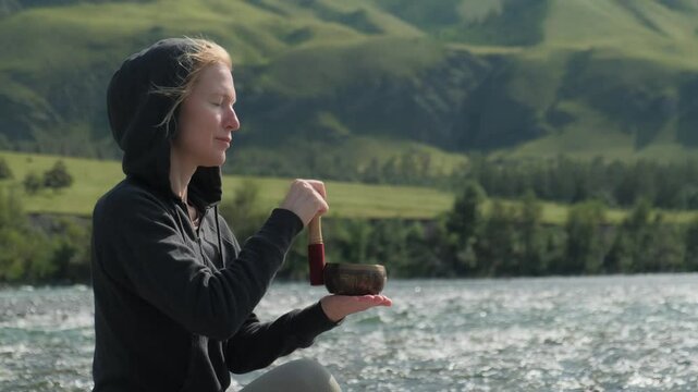Mindfulness woman drives a wooden stick over a singing tibetan bowl while sitting by the river in mountains. Zen atmosphere and sound meditation relieves stress.
