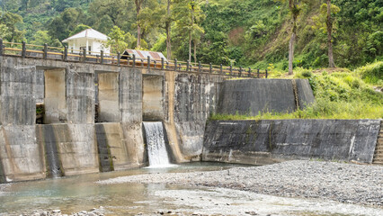A concrete dam with water flowing through the spillway, set in a lush rural landscape. A small house stands atop the structure, surrounded by greenery and hills.