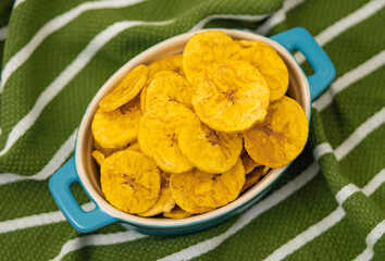 Crispy plantain chips in a blue dish on a green striped cloth background. Perfect for snack or tropical food concepts. © Octavio