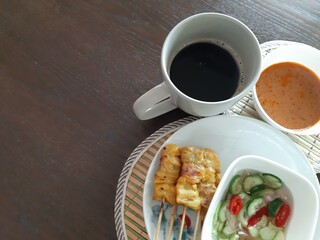 Top view of breakfast with pork satay with peanut sauce and pickles, which are cucumber slices and onions in vinegar with black coffee. Placed on an oak-colored table.