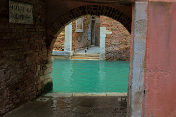 An arched passage frames a quiet canal with turquoise waters of Venice. A brick arch leads to a hidden corner with old walls and pigeons. Reflections on the wet stone add to the timeless charm.