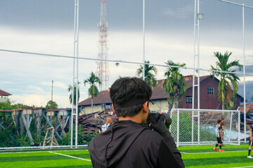 A photographer in a black outfit captures a penalty kick on a vibrant green soccer field, framed by a net fence and tropical palm trees, with rustic buildings in the background.