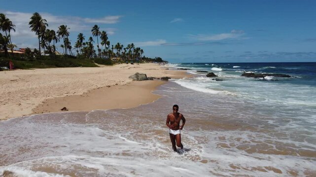 A Black Brazilian man in a white sunga walks toward the drone, waves, and jogs on a sunny Bahia beach. The drone ascends, unveiling a breathtaking view of the coastline and ocean.