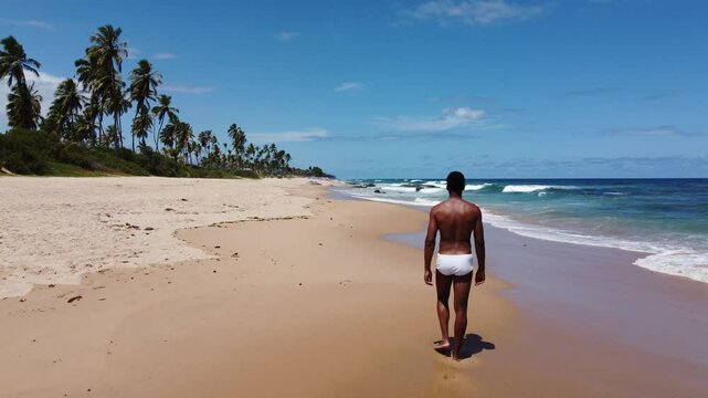 A Black Brazilian man in a white sunga walks calmly along the shore in Bahia. The drone ascends, unveiling a breathtaking coastline and turquoise waters.