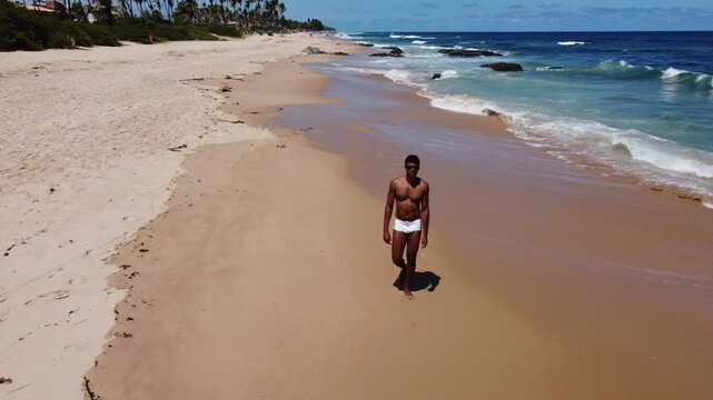 A Brazilian Black man in a white sunga walks along the Bahia beach shoreline, smiling before jogging on the sand, enjoying the sun and ocean breeze.