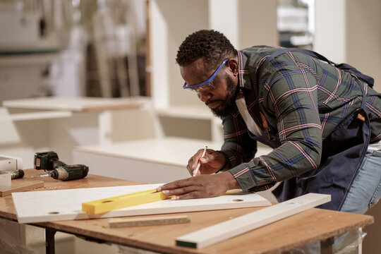 Affrican American male capenter use pensil and ruler working in furniture factory with concentrate, Black man labor work with piece of wood.