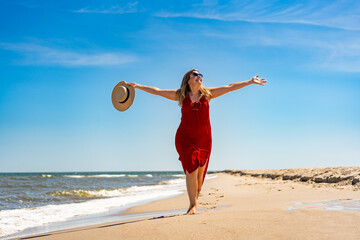 Beach holidays. Happy middle-aged woman wearing red dress with hat in raised up hand walking on...