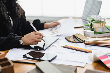 Architect uses tablet while reviewing interior drawing and material samples near a modern house model, Digital design and architectural planning. property development, building materials, 3d modeling.