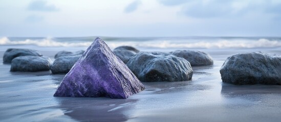 Purple triangular rock surrounded by smooth gray stones on a wet sandy beach with gentle ocean waves in the background.