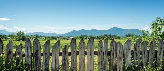 Fototapeta premium Rustic wooden fence framing a picturesque landscape in rural Thailand with mountains and clear blue skies in the background.
