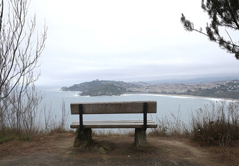 Photograph of a bench overlooking the sea in San Sebastiรกn, Spain, with a view of the city and waves on an overcast day. 