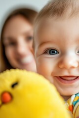 Baby smiling with a yellow plush toy, close-up, and a woman in the background, capturing joyful moments of parenthood and family bonding at home.