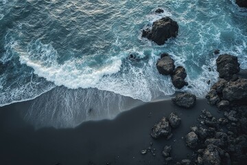 Aerial view of rugged coastline with dark sand, foamy waves, and jagged rocks under a cloudy sky
