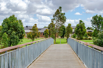 A wooden pedestrian bridge with metal railings in a landscaped park with lush green trees and modern residential houses in Australia. A suburban park with a boardwalk and outdoor space in neighborhood