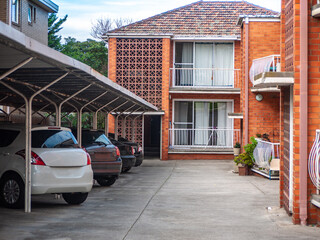 Covered parking area with parked cars sits adjacent to a two-story brick apartment building in an...