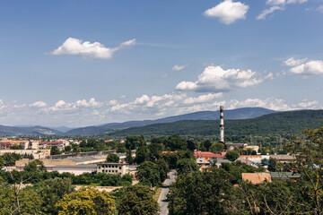 Obraz premium Rising above a small town nestled in a valley, an industrial chimney contrasts with lush greenery and rolling hills under a bright blue sky filled with fluffy clouds