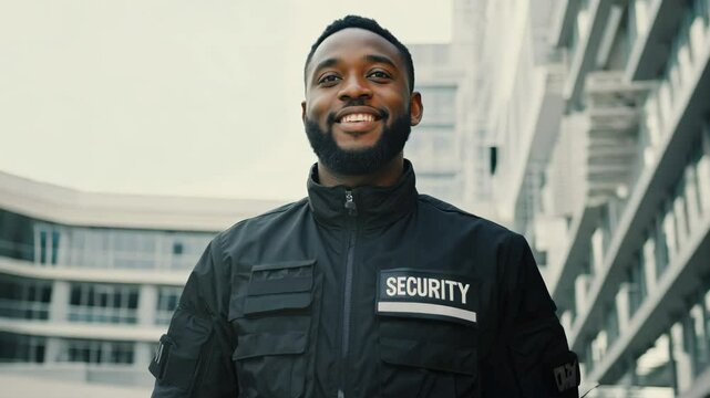 Young man security guard at night by modern office building in the city. Male security guard using portable radio transmitter near building outdoors. Wearing black security jacket.