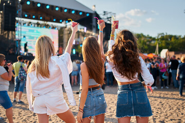 Back view of three women enjoying a music festival on sandy shore, raising drinks in cheers under...