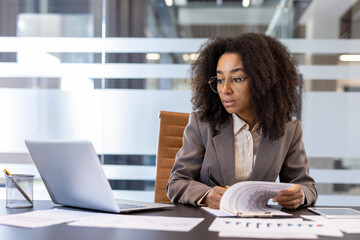 Serious young African American businesswoman working in office, sitting at workplace, looking at...