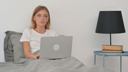 Serious Young Woman Looking at Camera while in Bed with Laptop