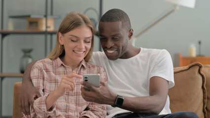 Interracial Couple Using Phone while Sitting on Sofa at Home