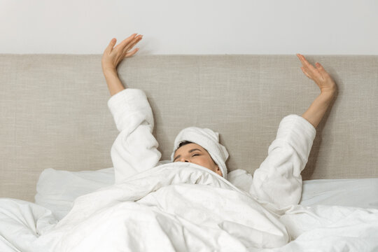 woman in bedroom with towel on head and in bathrobe waking up in the morning,outstretching hand,rub eyes.beautiful girl drinking tea or coffee,sitting in hands with cup.female lying on bed, relaxing.