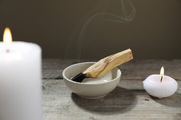 Smoldering palo santo stick and burning candles on wooden table against grey background, closeup